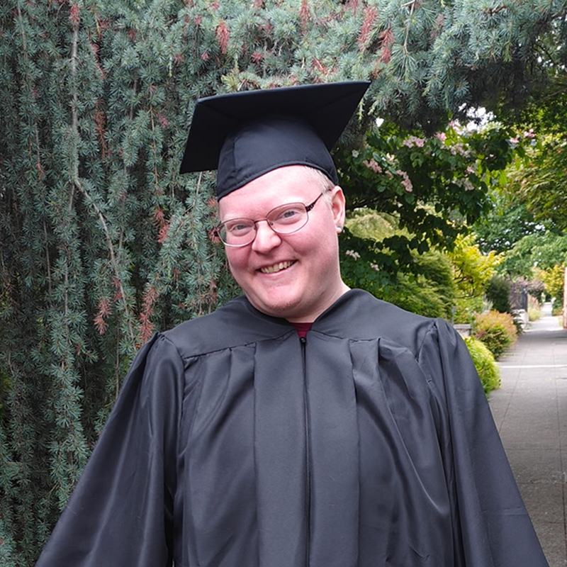Photo of Rory Hackney, a white person in a black graduation cap and gown, smiling in front of some pine trees.